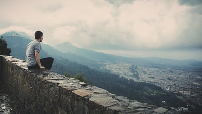 在多云的天空下，一个人独自坐在面向山城的水泥砖墙上